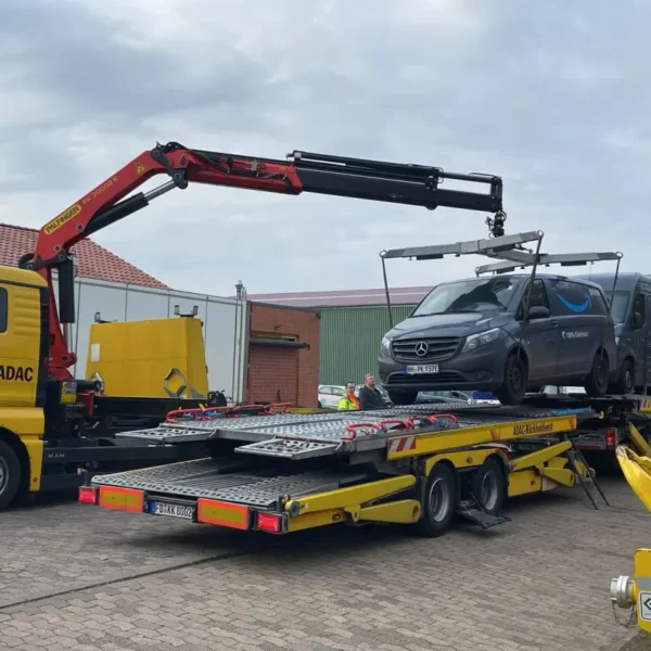 Gelber ADAC-Lkw mit Kran hebt einen grauen Mercedes-Transporter auf einen Autotransporter-Anhänger. Die Szene zeigt einen professionellen Fahrzeugumschlag auf einem Betriebshof mit mehreren Personen und Werkstattausrüstung im Hintergrund.