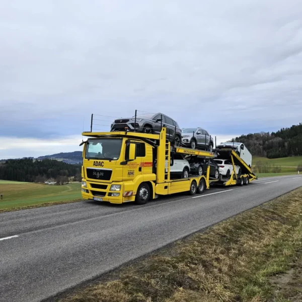 Gelber ADAC-Autotransporter mit beladenen Fahrzeugen auf einer ruhigen Landstraße in ländlicher Umgebung. Das Fahrzeug gehört zum Rückholdienst und fährt durch hügelige Landschaft mit Feldern und Wäldern unter bewölktem Himmel.
