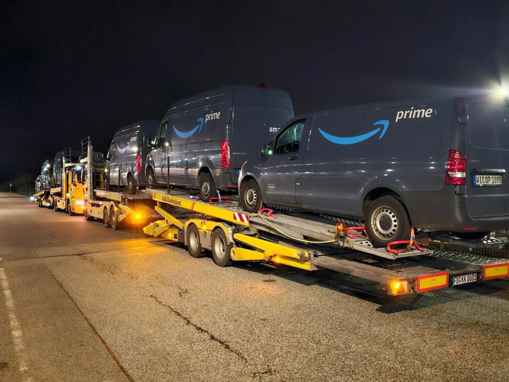 Gelber ADAC-Autotransporter beladen mit mehreren Amazon Prime Lieferwagen bei Nacht auf einem Parkplatz. Das Fahrzeug des Rückholdienstes steht unter künstlicher Beleuchtung, bereit für den Weitertransport.
