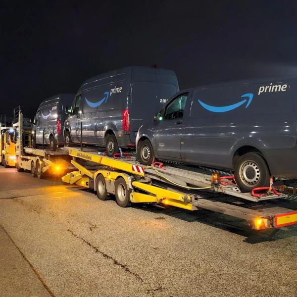 Gelber ADAC-Autotransporter beladen mit mehreren Amazon Prime Lieferwagen bei Nacht auf einem Parkplatz. Das Fahrzeug des Rückholdienstes steht unter künstlicher Beleuchtung, bereit für den Weitertransport.
