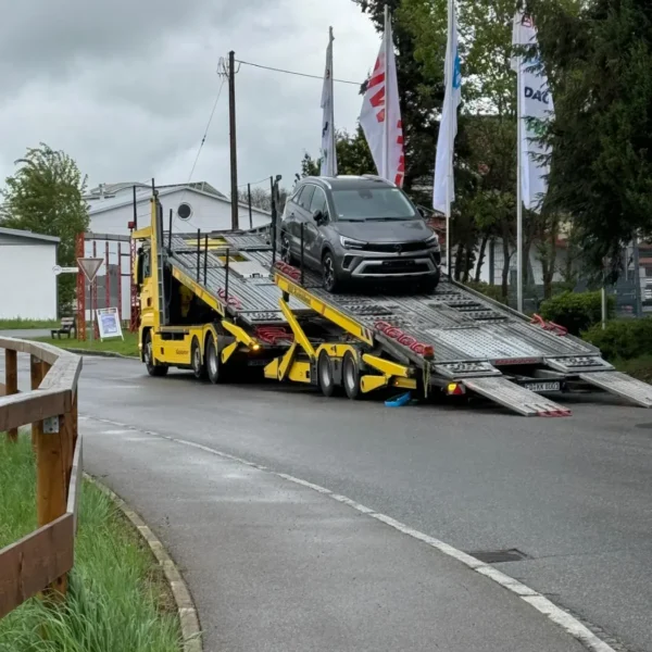 Ein Fahrzeug wird auf einen gelben ADAC-Autotransporter verladen. Der Lkw steht auf einer regennassen Straße vor einem Gebäude mit mehreren Fahnen. Die Szene zeigt den professionellen Ablauf der Fahrzeuglogistik im Rückholdienst.