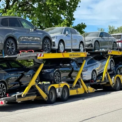 Gelber ADAC-Autotransporter mit mehreren hochwertigen Audi-Fahrzeugen, geparkt bei sonnigem Wetter unter grünen Bäumen. Die Fahrzeuge sind auf zwei Ebenen verladen, sichtbar ist der Rückholdienst im Einsatz.