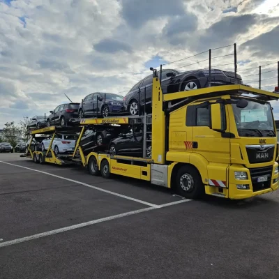 Gelber ADAC-Autotransporter vom Rückholdienst steht auf einem markierten Parkplatz, beladen mit verschiedenen Neuwagen. Der Himmel ist leicht bewölkt, das Bild vermittelt ein professionelles Logistik-Szenario bei Tageslicht.